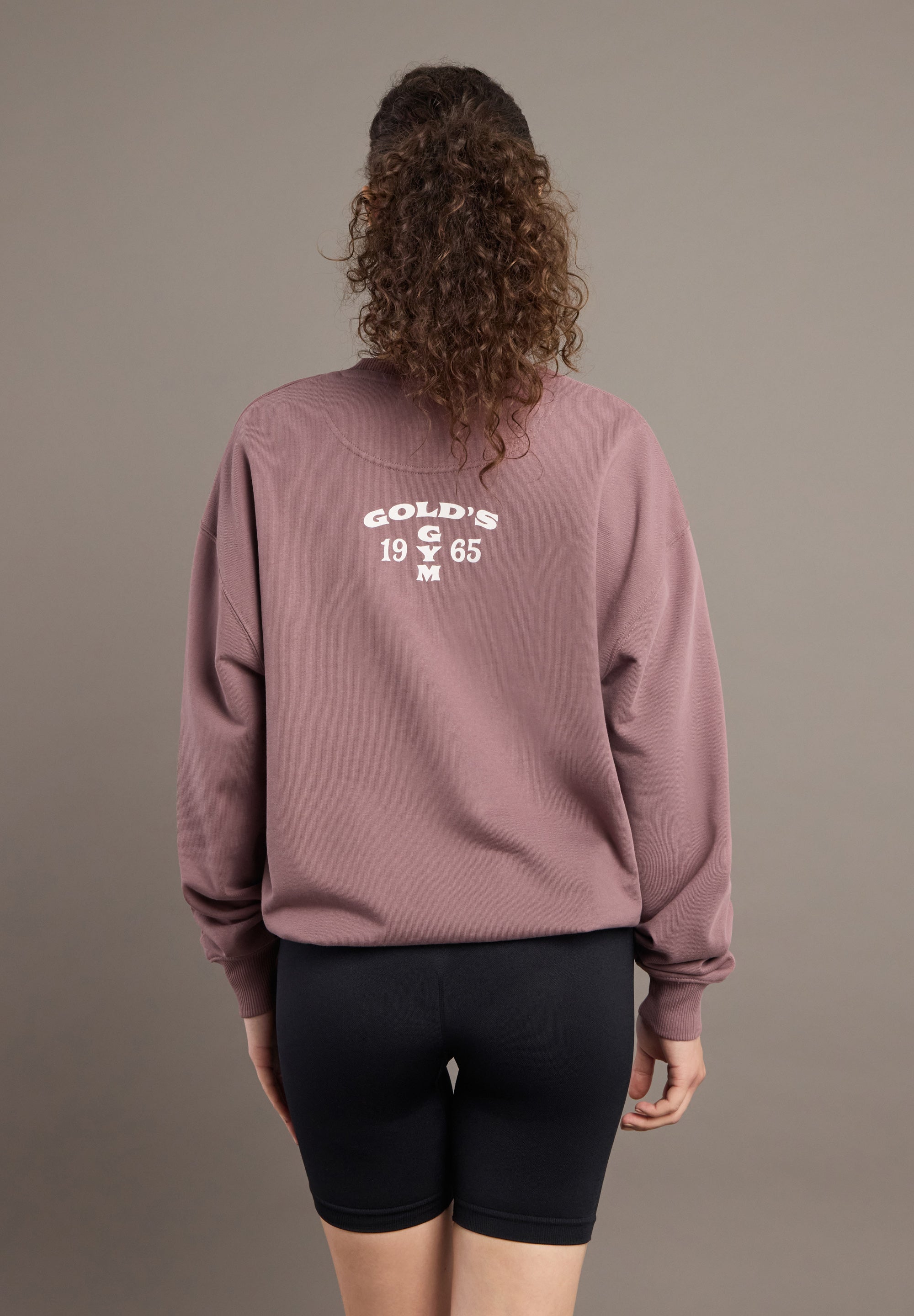 Person wearing a mauve sweatshirt with text on the back against a brown background