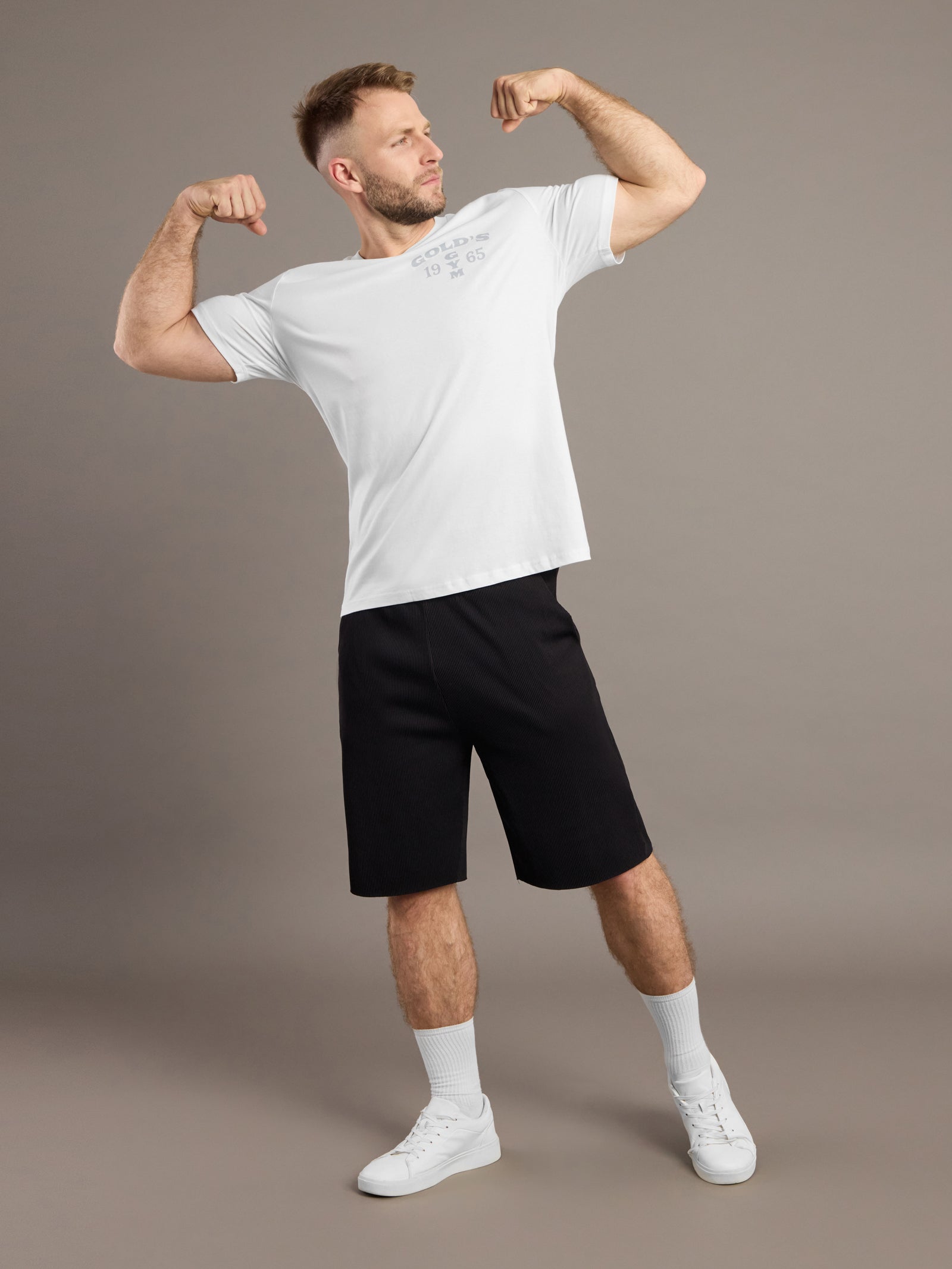 Man flexing muscles wearing a white t-shirt and black shorts on a brown background