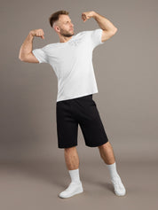 Man flexing muscles wearing a white t-shirt and black shorts on a brown background
