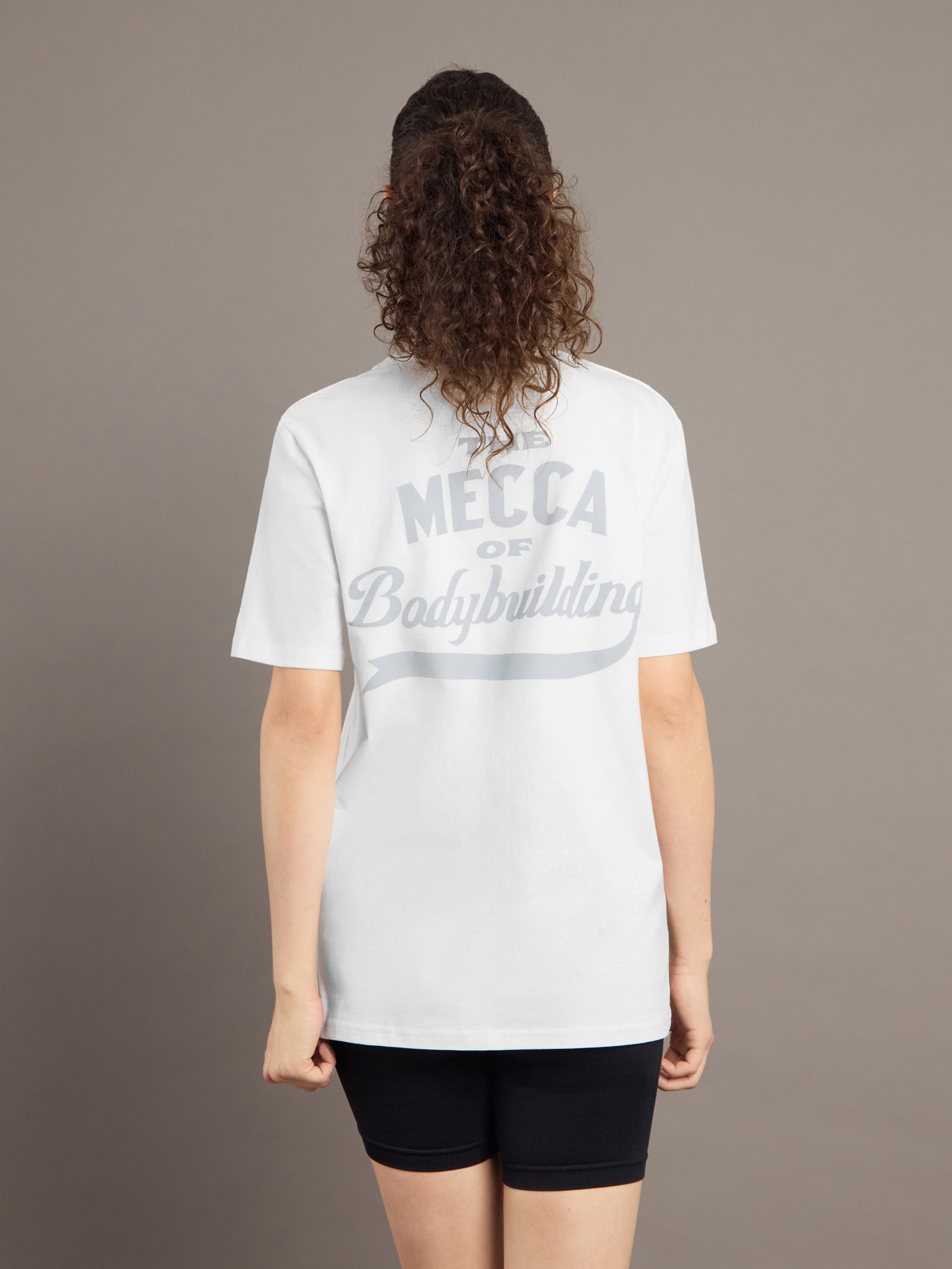 Person wearing a white t-shirt with 'Mecca of Bodybuilding' text on the back against a brown background