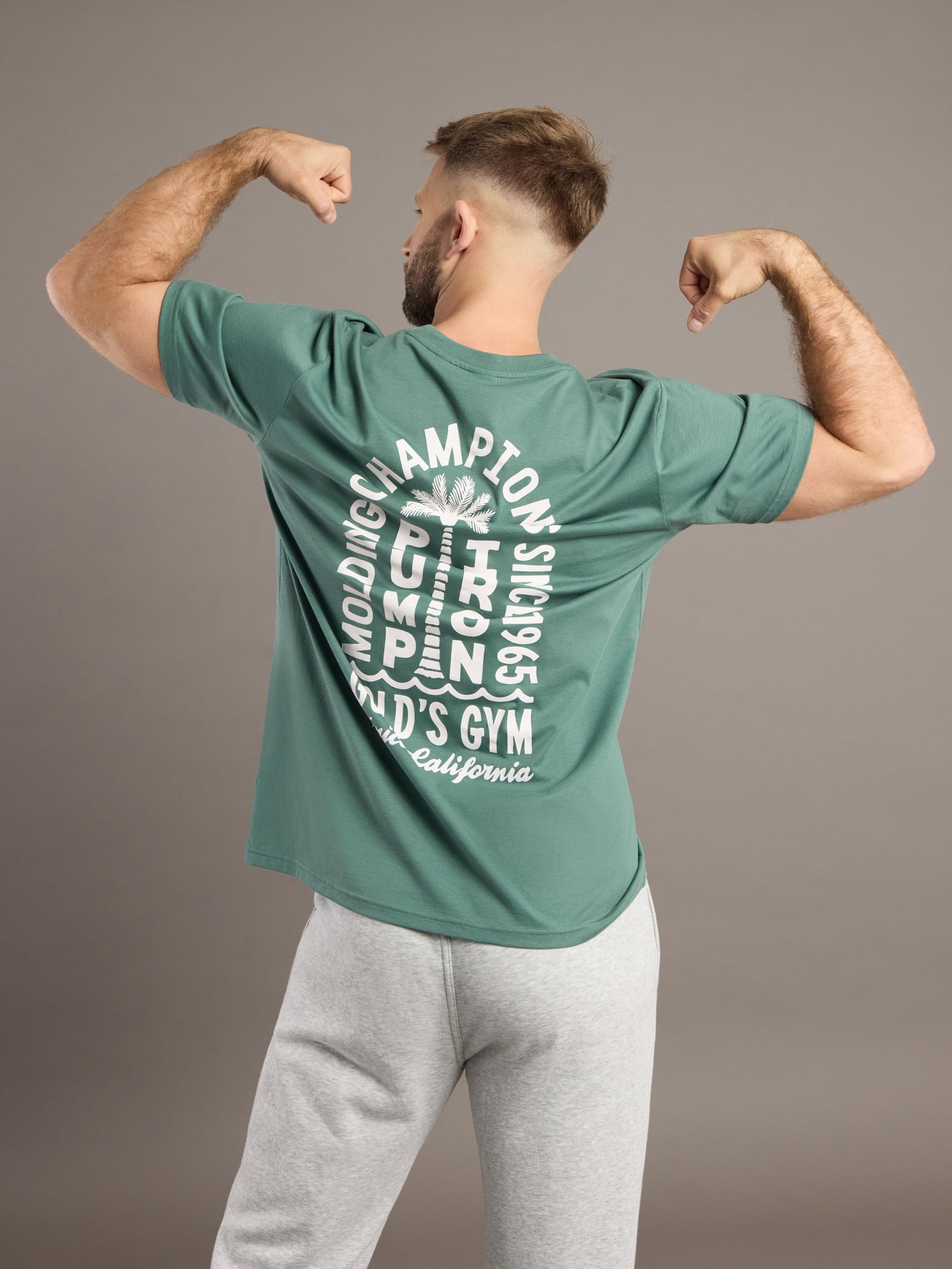 Man wearing a green t-shirt with text on the back, flexing his muscles against a brown background