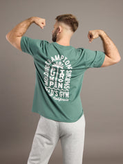 Man wearing a green t-shirt with text on the back, flexing his muscles against a brown background