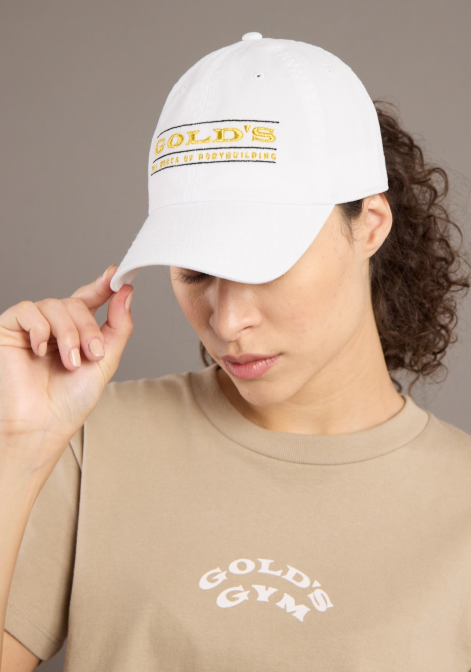 Person wearing a white cap with 'Gold's' branding and a beige t-shirt with 'Gold's Gym' branding against a neutral background.