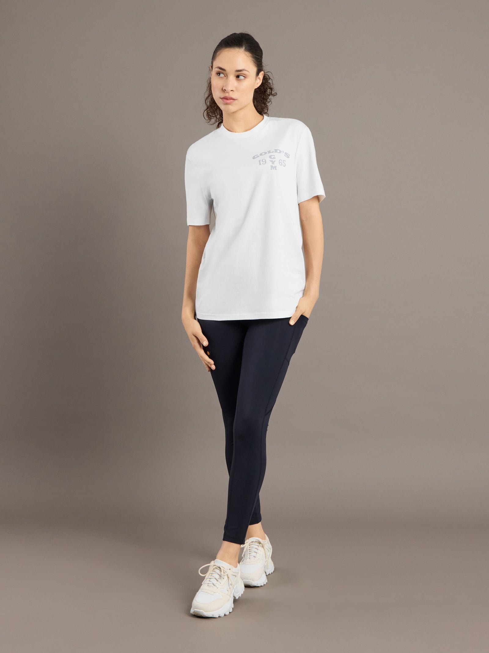 Woman wearing a white t-shirt with a logo and black leggings on a brown background