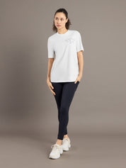 Woman wearing a white t-shirt with a logo and black leggings on a brown background