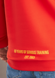 Red sweatshirt with yellow text '60 YEARS OF SERIOUS TRAINING EST. 1965' on a gray background