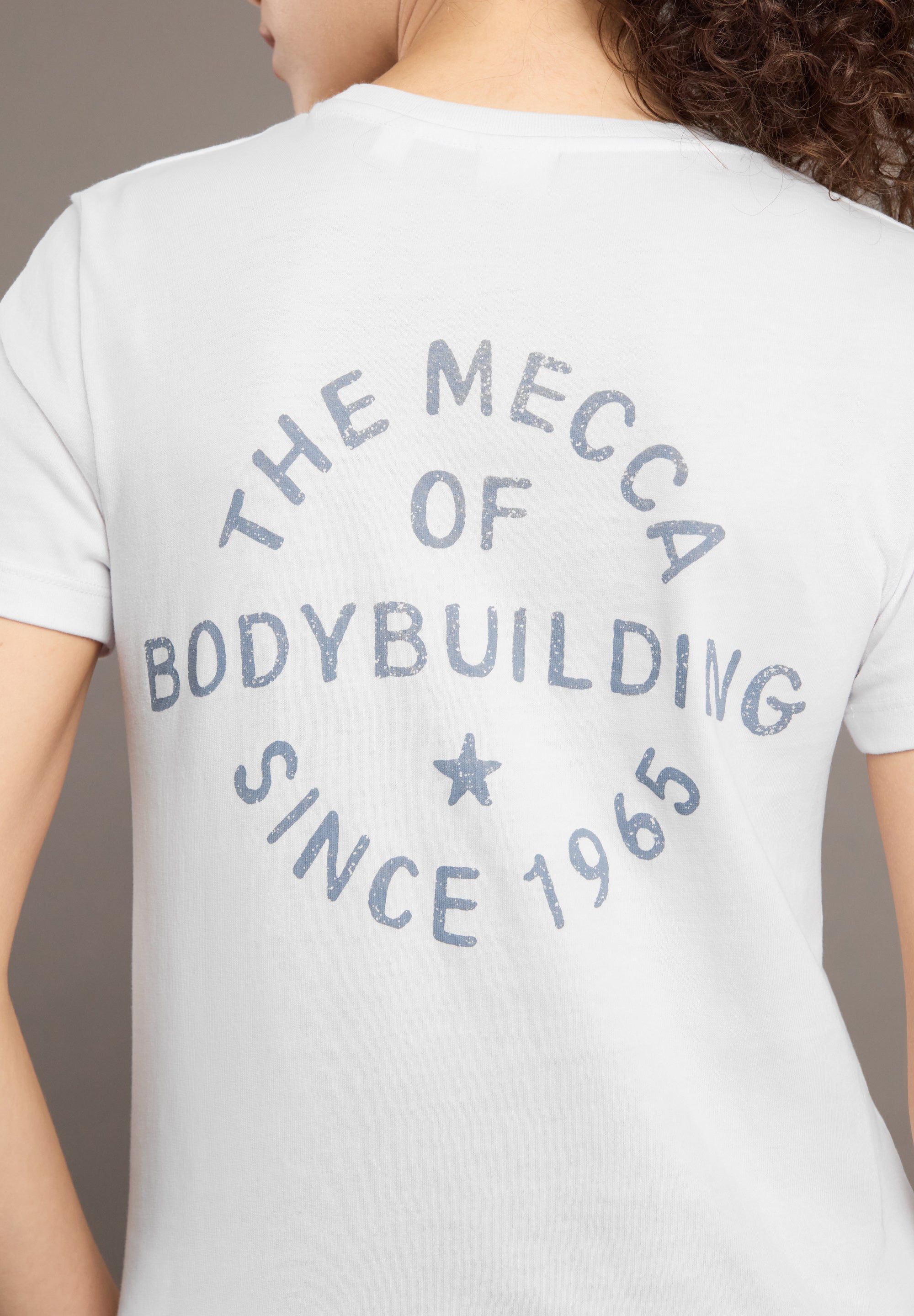 Person wearing a white t-shirt with 'The Mecca of Bodybuilding Since 1965' text.