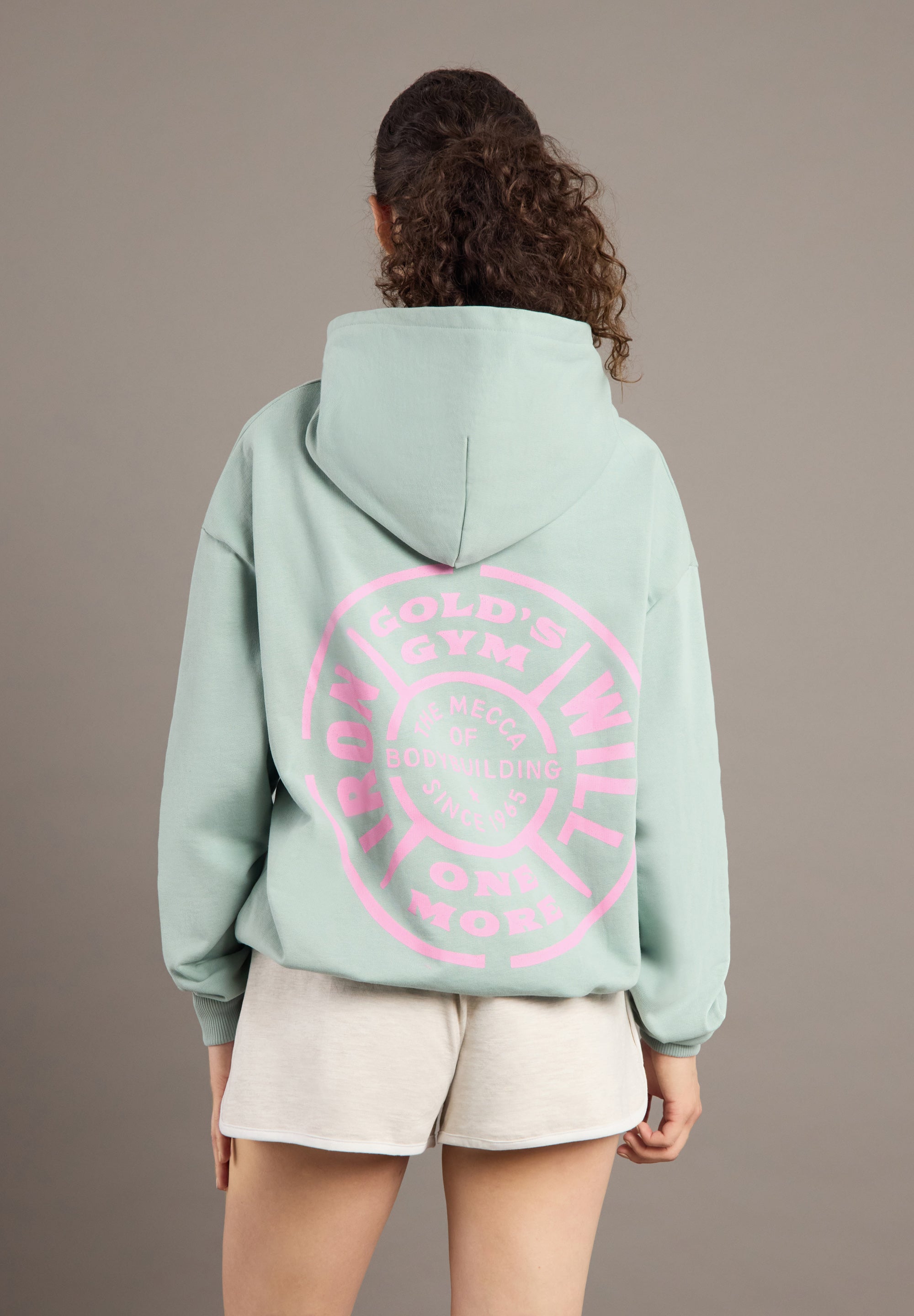 Person wearing a light green hoodie with pink text on the back against a plain background