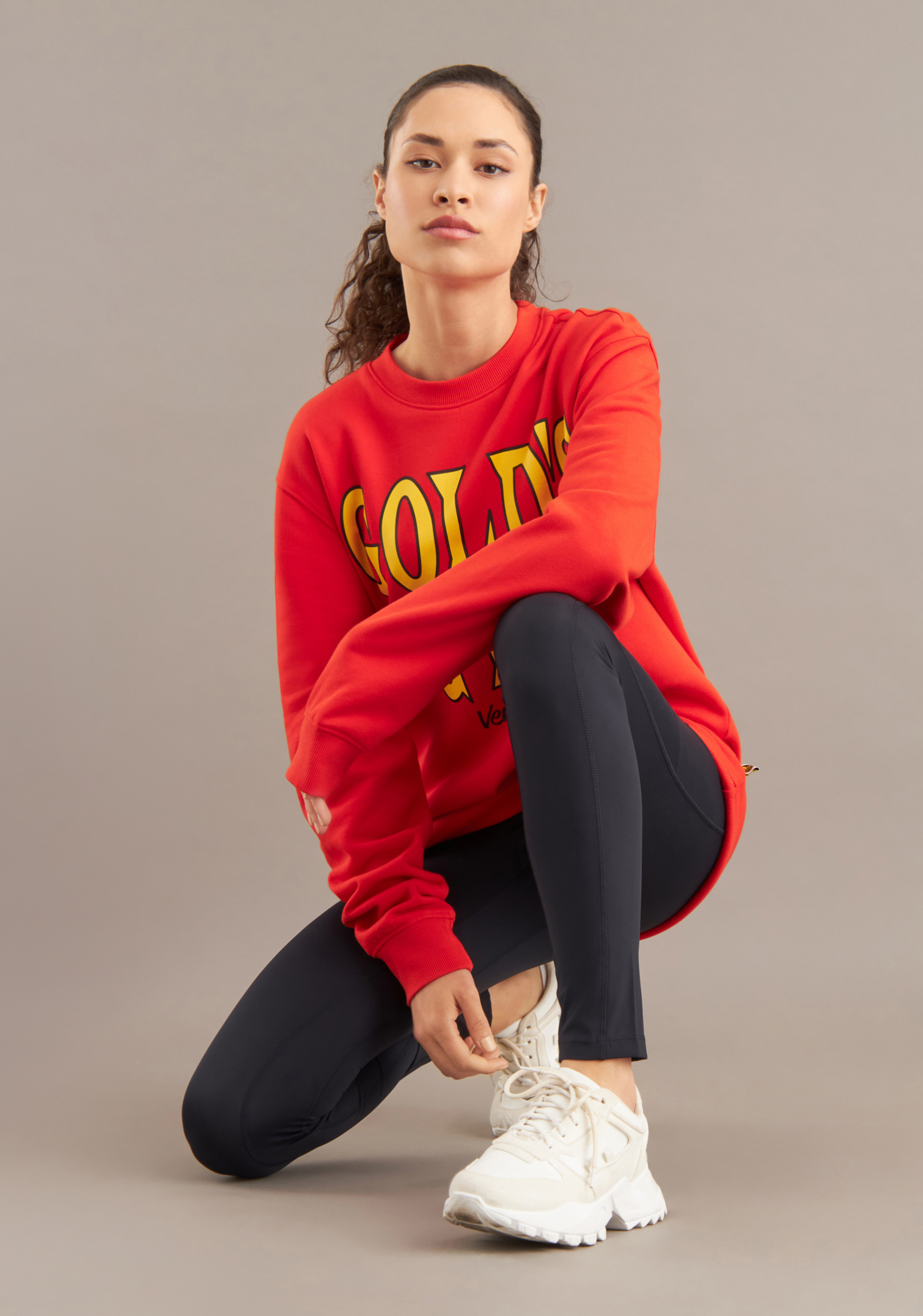 Person wearing a red sweatshirt with yellow text, black pants, and white sneakers on a beige background