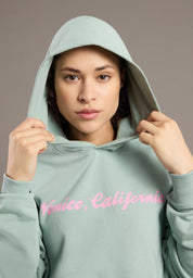 Person wearing a light green hoodie with 'Venice, California' text in pink on a neutral background