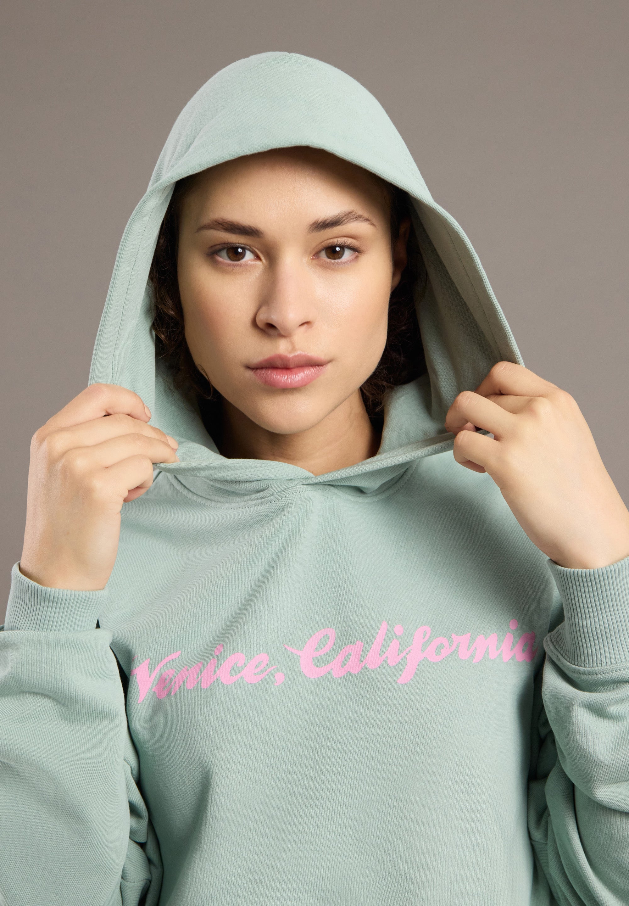 Person wearing a light green hoodie with 'Venice, California' text in pink on a neutral background