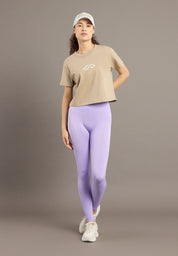 Woman wearing a beige t-shirt, purple leggings, and a white cap on a brown background