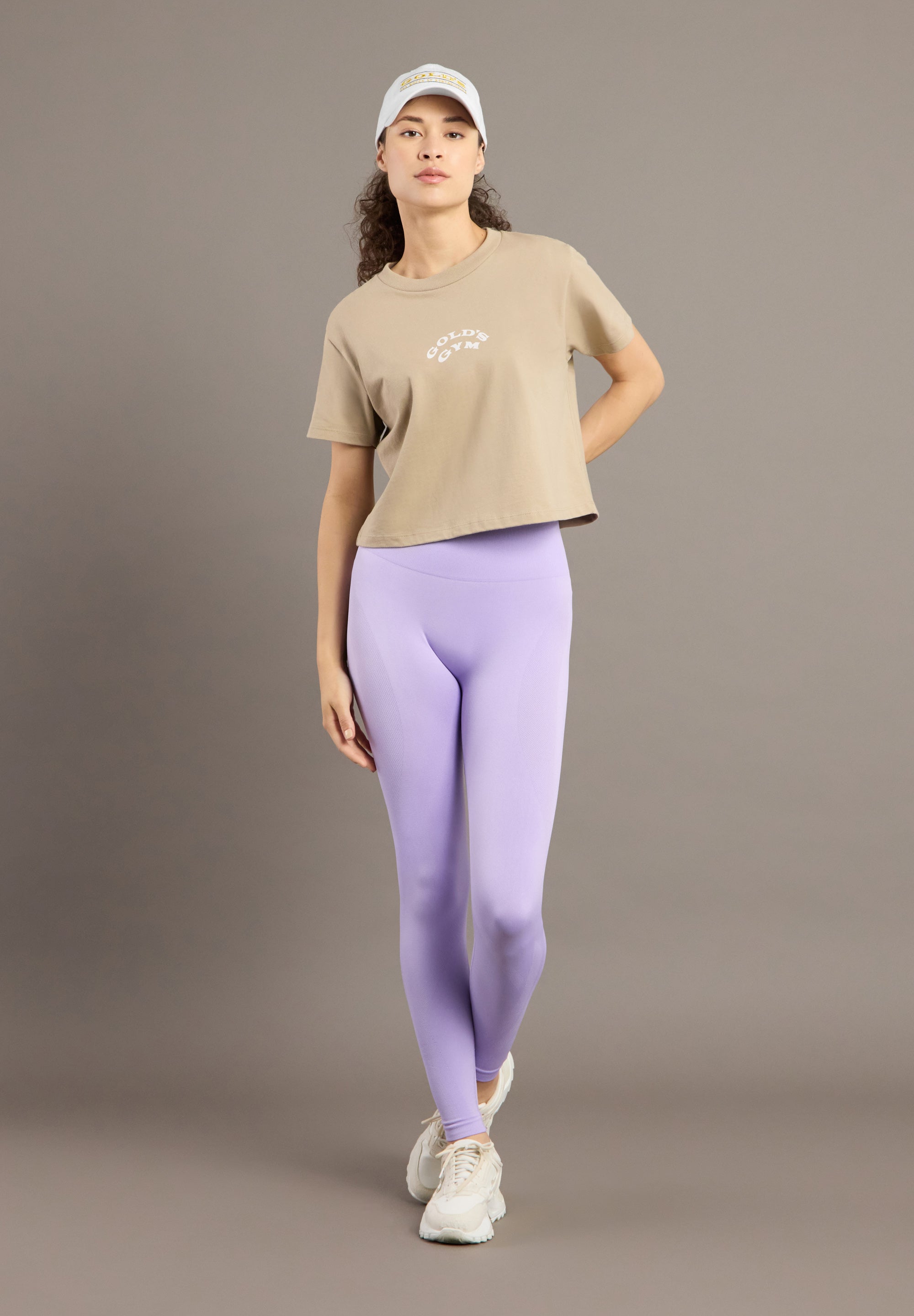 Woman wearing a beige t-shirt, purple leggings, and a white cap on a brown background