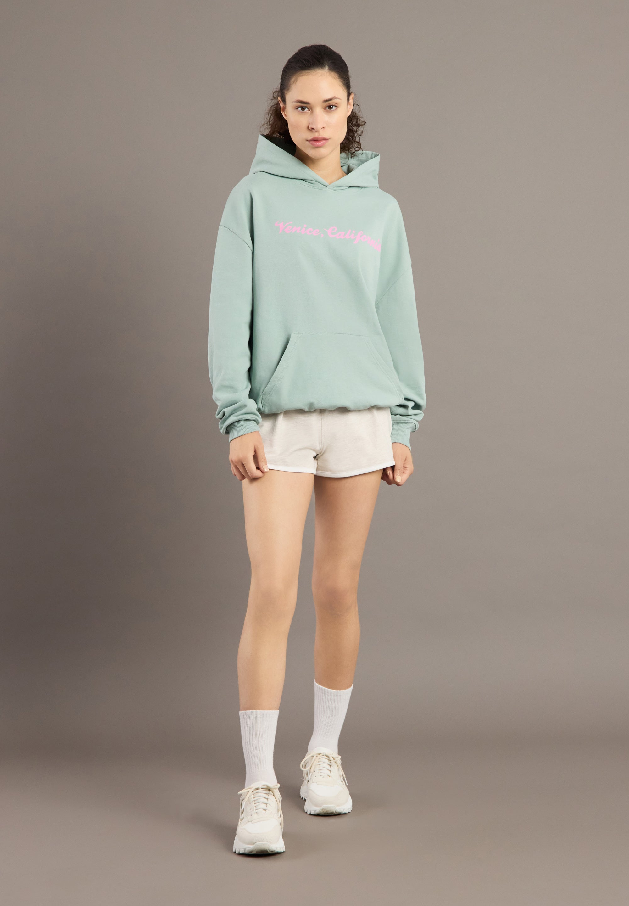 Person wearing a light green hoodie with pink text, white shorts, and white sneakers on a brown background