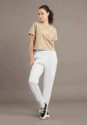 Woman wearing a beige t-shirt and light gray pants against a brown background