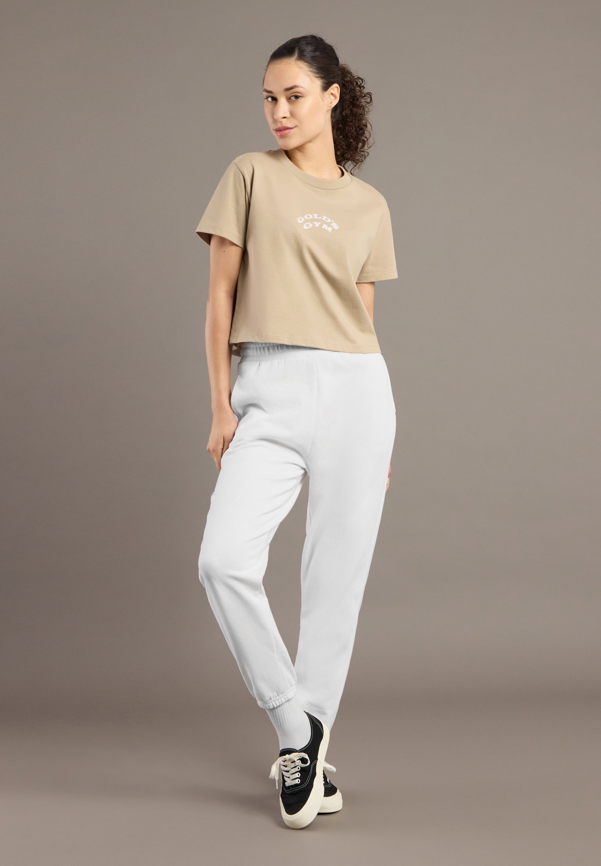 Woman wearing a beige t-shirt and light gray pants against a brown background